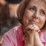 Senior woman with short brunette hair and pink shirt, hands clasped, looking thoughtful outdoors.